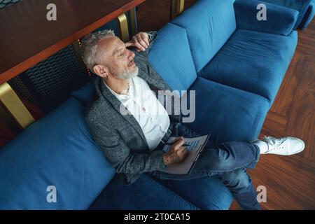 Imprenditore anziano penoso che tiene appunti mentre si siede sul divano nel coworking Foto Stock