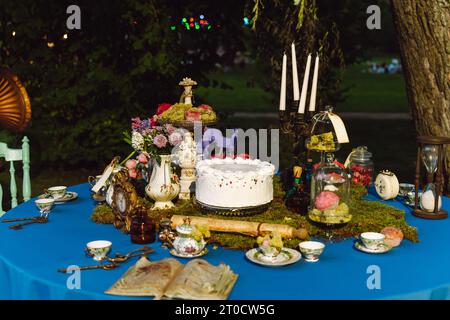 Il tavolo da pranzo e' decorato nello stile di Alice nel Paese delle meraviglie. Un vecchio libro aperto, una torta, ciotole da tè, candele in un candelabro, una clessidra su un Foto Stock