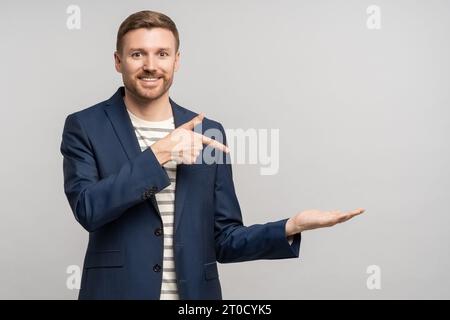 Un uomo d'affari sorridente e barbuto che punta il dito indice a portata di mano sullo spazio di copia su sfondo grigio. Foto Stock