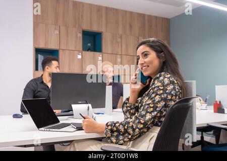 Donna felice che parla con il cellulare mentre lavora nell'ufficio di una collaborazione accanto ai colleghi Foto Stock