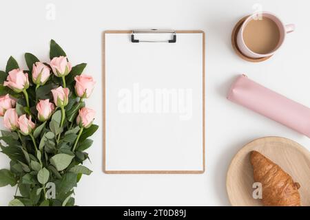 Vista dall'alto di un mockup in legno con un bouquet di rose rosa, caffè e croissant su un tavolo bianco. Foto Stock