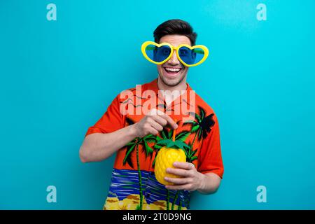 Ritratto di un ragazzo positivo e amichevole con setole. Indossa una camicia tropicale con cuori, indossa una tazza di ananas isolata su sfondo blu Foto Stock