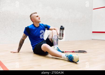 Giovane uomo in abbigliamento sportivo seduto sul pavimento, riposante dopo l'allenamento di squash sul campo da squash. Sudorazione, acqua potabile Foto Stock