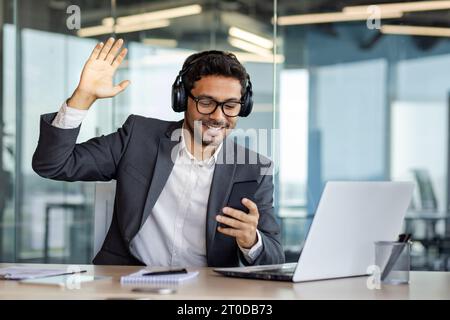 Giovane uomo d'affari gioioso che si rilassa in ufficio, uomo in cuffia ascolta musica, canta e balla, utilizza l'app sul telefono, lavora con il computer portatile con lo smartphone in mano. Foto Stock