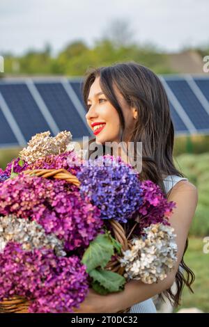 Bella giovane bruna donna caucasica sorridente con mazzi di fiori lilla, primo piano. Pannelli solari sullo sfondo. Foto Stock