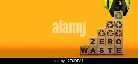 Guanti disposti a mano in blocchi di legno con testo Zero rifiuti e simboli di riciclaggio su sfondo arancione e giallo con spazio per la copia. Foto Stock
