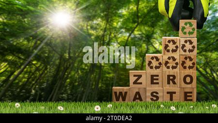 Guanti disposti a mano in blocchi di legno con testo Zero rifiuti e simboli di riciclaggio su un prato verde con fiori e foresta verde sullo sfondo. Foto Stock