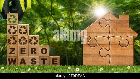 Guanti disposti a mano in blocchi di legno con testo Zero Waste e simboli di riciclaggio su un prato verde con una piccola casa in legno fatta di pezzi puzzle. Foto Stock