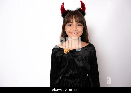 halloween, festività e infanzia: una ragazza sorridente in costume nero e corna rosse del diavolo su sfondo bianco Foto Stock