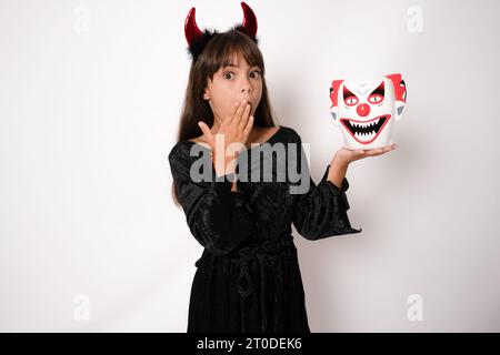 halloween, festività e infanzia: una ragazza sorridente in costume nero e corna rosse del diavolo su sfondo bianco Foto Stock