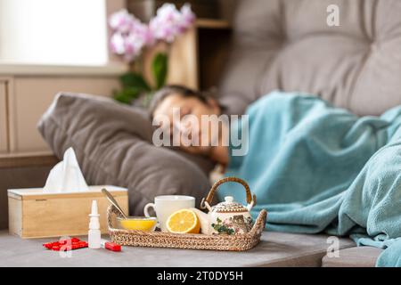 Epidemia di influenza. Una donna malata dorme sotto una coperta sul divano. Davanti a lei c'è un vassoio con una teiera, limone e miele. Nelle vicinanze ci sono le pillole, un The Foto Stock