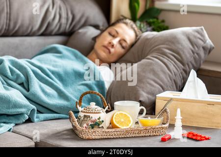 Epidemia di influenza. Una donna malata dorme sotto una coperta sul divano. Davanti a lei c'è un vassoio con una teiera, limone e miele. Nelle vicinanze ci sono le pillole, un The Foto Stock