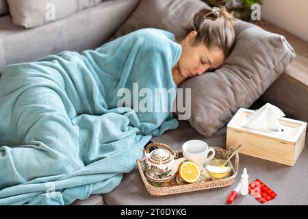 Epidemia di influenza. Una donna malata dorme sotto una coperta sul divano. Davanti a lei c'è un vassoio con una teiera, limone e miele. Nelle vicinanze ci sono le pillole, un The Foto Stock