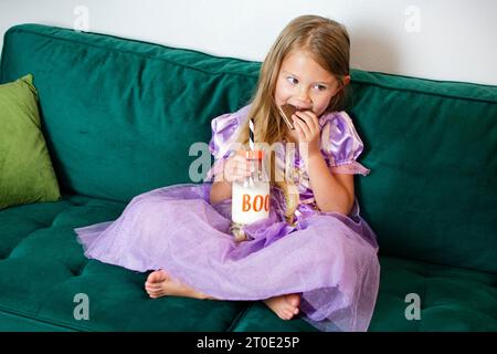 La graziosa bambina caucasica sta mangiando biscotti in costume di Halloween. Ragazza con vestito viola. Concetto bambini e Halloween Foto Stock