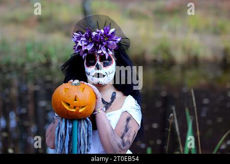 Una ragazza seria con uno spaventoso outfit da sposa morta, con lanterna Jack o zucca che guarda la fotocamera mentre si trova nella natura durante le vacanze di Halloween Foto Stock