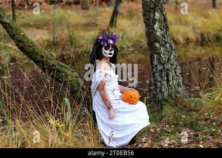 Una ragazza seria con uno spaventoso outfit da sposa morta, con lanterna Jack o zucca che guarda la fotocamera mentre si trova nella natura durante le vacanze di Halloween Foto Stock