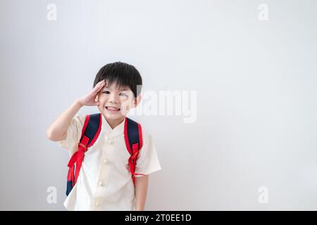 Ragazzo asiatico che indossa abiti tradizionali tailandesi, in piedi con una borsa della scuola. Fingere di salutare e prepararsi per la scuola su sfondo bianco Foto Stock