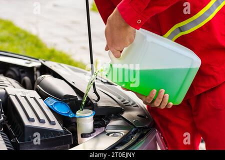 Operatore addetto alla manutenzione da vicino, conducente che versa liquido lavavetri per la pulizia del parabrezza antigelo nel serbatoio dell'auto del cofano dalla bottiglia del filtro a carboni attivi. Detergente liquido per vetri per parabrezza per uso estivo o invernale Foto Stock