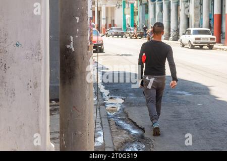 Un uomo cubano cammina lungo il marciapiede dove si vede acqua sporca e stagnante. I tradizionali vecchi edifici con le intemperie sono sullo sfondo dello stile della strada Foto Stock