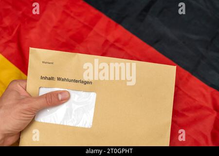 Augusta, Baviera, Germania - 29 settembre 2023: Mano con in mano una busta con i documenti elettorali per le elezioni statali davanti a una bandiera tedesca *** Hand hält einen Umschlag mit Wahlunterlagen zur Landtagswahl vor eine Deutschland Flagge Foto Stock