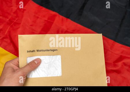 Augusta, Baviera, Germania - 29 settembre 2023: Mano con in mano una busta con i documenti elettorali per le elezioni statali davanti a una bandiera tedesca *** Hand hält einen Umschlag mit Wahlunterlagen zur Landtagswahl vor eine Deutschland Flagge Foto Stock