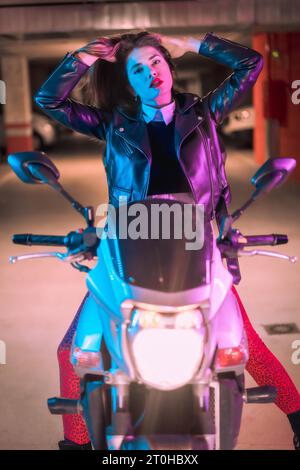 Fotografia con neon blu e rosa su una motocicletta. Ritratto di una giovane modella bionda caucasica che indossa una giacca di pelle nera Foto Stock