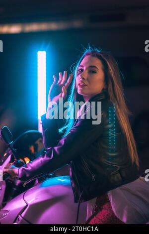 Fotografia con neon blu e rosa su una moto da dietro. Ritratto di una giovane modella bionda caucasica che indossa una giacca di pelle nera Foto Stock