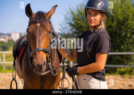 Posa di una ragazza bionda caucasica su un cavallo che accarezza e coccola un cavallo marrone, vestito di cavaliere nero con berretto di sicurezza Foto Stock