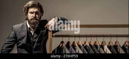 Sarto, sartoriale. Elegante abito da uomo. Bellissimo uomo di moda barbuto in costume classico. Abiti maschili appesi di fila. Vestito da uomo, sarto nel suo Foto Stock
