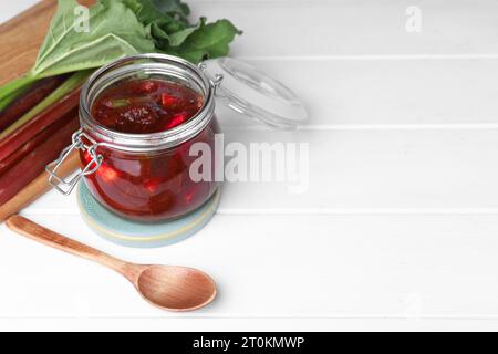 Barattolo di gustosa marmellata di rabarbaro, steli e cucchiaio su un tavolo di legno bianco. Spazio per il testo Foto Stock