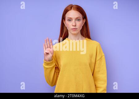 bella donna seria sicura di sé rossa in elegante maglione giallo che mostra il simbolo del linguaggio dei segni a 4 numeri per gli umani sordi con sfondo blu. io Foto Stock