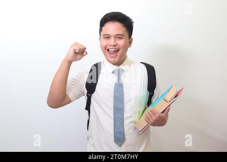 Studente delle superiori indonesiane che indossa un'uniforme bianca con cravatta grigia che tiene alcuni libri, sorridendo e alzando il pugno, celebrando il successo. Foto Stock