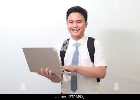 Studente delle superiori indonesiane che indossa un'uniforme bianca con una cravatta grigia che regge un portatile, su sfondo grigio Foto Stock