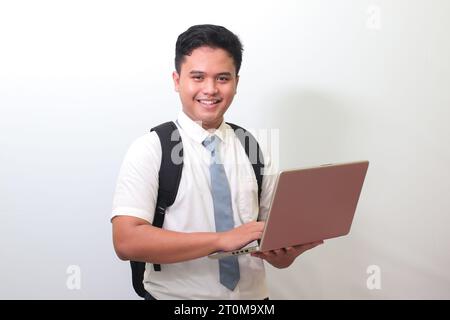 Studente delle superiori indonesiane che indossa un'uniforme bianca con una cravatta grigia che regge un portatile, su sfondo grigio Foto Stock