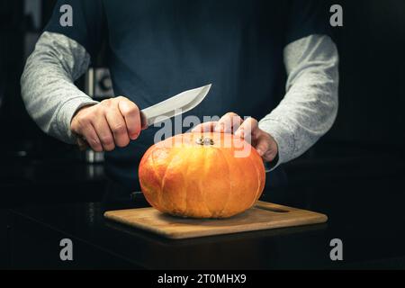 Primo piano delle mani dell'uomo che intagliano una piccola zucca arancione nel jack-o-lantern per la decorazione natalizia di Halloween con il coltello. Immagine con messa a fuoco selettiva Foto Stock