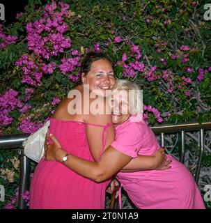 Madre e figlia con abiti rossi accesi si si abbracciano nei pressi di bouganvillea Bush di notte 5th Centennial Avenue, Puerto de Santiago, Tenerife. Foto Stock