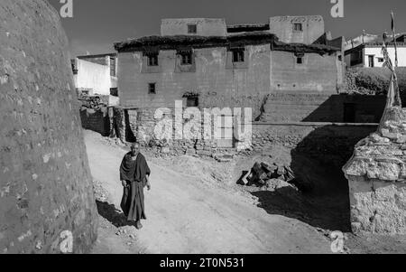 Il monaco buddista in abiti tradizionali cammina lungo una strada sterrata fiancheggiata da case nel villaggio di Kibber, Himachal Pradesh, India. Foto Stock