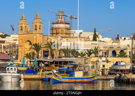 Marsaxlokk, Malta - 14 ottobre 2019 - Porto e skyline di Marsaxlokk tradizionale villaggio di pescatori con barca luzzu e Chiesa Parrocchiale di nostra Signora di Pom Foto Stock