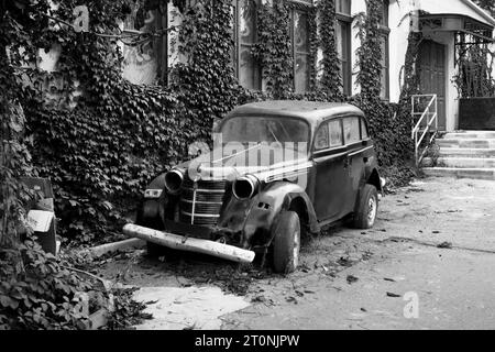 Foto in bianco e nero di un'auto retrò in piedi vicino alla casa. Vecchia macchina arrugginita. Foto Stock