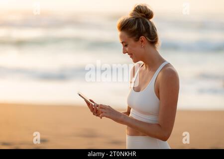 Allegra atleta europea in abbigliamento sportivo, con cellulare, in piedi sulla spiaggia di mare dopo l'allenamento all'aperto Foto Stock