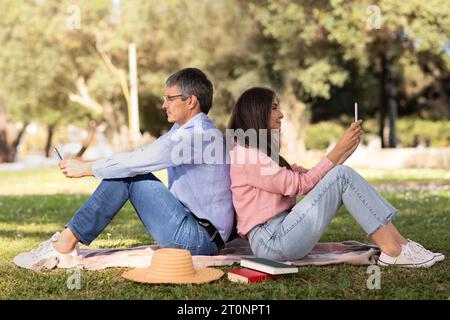 Coppia matura seduta da una parte all'altra e che usa gli smartphone a Park Outdoors Foto Stock