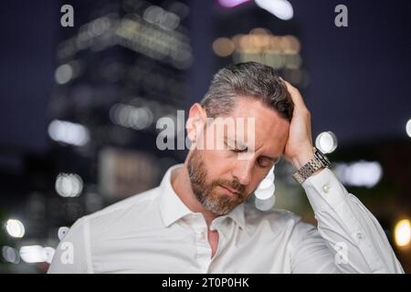 Ritratto ravvicinato di un uomo d'affari stressato sullo sfondo notturno della città. Il ceo manager stressato ha stress e mal di testa emicrania all'aperto. Stanco freelance Foto Stock