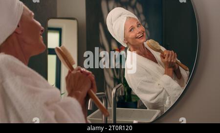 Spensierata donna anziana caucasica matura in accappatoio dopo la doccia sorridente signora di mezza età nel bagno di casa canta al punto di ballo del microfono della spazzola Foto Stock