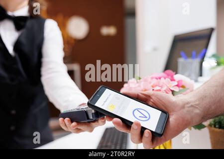 Vista dettagliata del concierge che aiuta i toruisti anziani con i pagamenti tramite smartphone e tecnologia NFC. Primo piano di un viaggiatore di sesso maschile che paga tramite dispositivo mobile con l'assistenza dell'addetto alla reception dell'hotel. Foto Stock