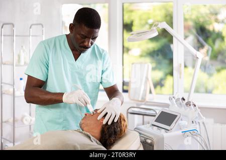 Cosmetologo afroamericano che esegue la procedura di radiofrequenza facciale sull'uomo Foto Stock