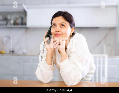 Giovane donna che si appoggia al tavolo in cucina, prende lo smartphone Foto Stock