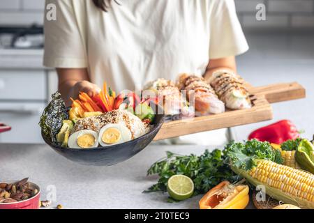 Recipiente con verdure, riso e involtini primavera in mani femminili in cucina. Foto Stock