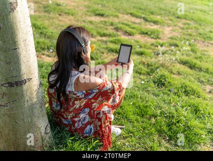 Donna con cuffie che legge un e-book con schermo vuoto mentre è seduta all'aperto sull'erba. Concetto tecnologico. Foto Stock