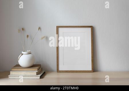 Mockup verticale con cornice in legno vuota sul tavolo. Moderno vaso rotondo in ceramica con coda di coniglio asciutta, erba di Lagurus ovatus e vecchi libri. Muro bianco Foto Stock