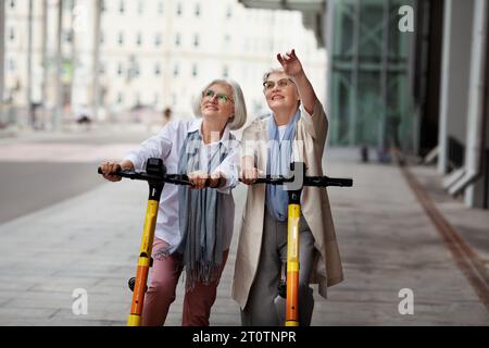 Ritrarre donne anziane soddisfatte con i capelli grigi che si divertono a cavalcare insieme su scooter elettrici all'aperto. Le amiche anziane esplorano la città in un'escursione, Foto Stock